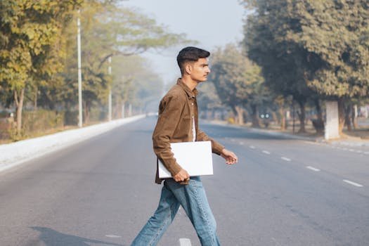 Young adult walking across a tree-lined street holding a laptop, symbolizing modern urban life.