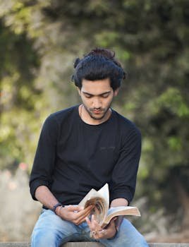 A young man in casual clothing sits outdoors, engrossed in reading a book.